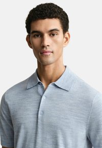 Polo bleu clair en tissu texturé, avec un col classique et deux boutons sur le devant, arborant un logo discret sur la poitrine.