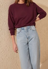 Donna che indossa una camicia a maniche lunghe bordeaux infilata in jeans a vita alta di un blu chiaro, in piedi contro un muro beige testurizzato.