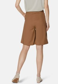 Bruine op maat gemaakte shorts met een gladde textuur, knielang design en subtiele zijzakken. Gepaard met lichtbeige schoenen.