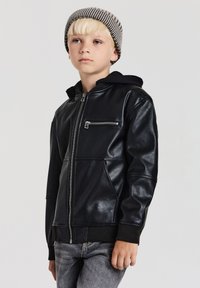 Bambino biondo che indossa un berretto a righe, una giacca in pelle nera con zip e tasca sul petto, e jeans grigi, in piedi contro uno sfondo semplice.