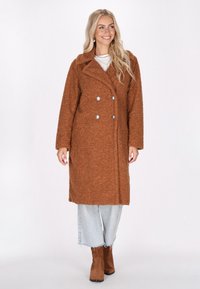 Brun texturerad lång kappa med dubbelknäppt design, stora slag och silverknappar, parat med ljus denim och bruna ankelstövlar.