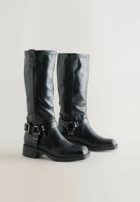 Mustad nahast kuni põlveni ulatuvad saapad, millel on sile tekstuur, pandla detailid ja madal plokkkonts. Üksikasjadeks on ülemises osas mugavaks selga tõmbamiseks mõeldud tõmblused.