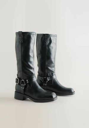 Czarne skórzane kozaki do kolan o gładkiej teksturze, z akcentami w postaci klamer i niskim słupkowym obcasem. Posiadają pętle u góry ułatwiające zakładanie.