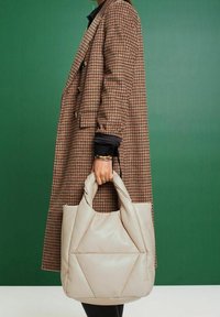 Beige polstret mulepose med geometrisk design, der har to rundede håndtag og en blød tekstur. Bæres sammen med en mønstret frakke.