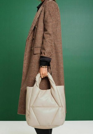 Beige vadderad toteväska med geometrisk design, försedd med två runda handtag och en mjuk textur. Använd tillsammans med en mönstrad kappa.