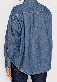 Chemise en denim bleu moyen, coupe décontractée avec empiècement au dos, manches longues, poignets à boutons et ourlet arrondi. Texture douce avec des détails de couture minimalistes.