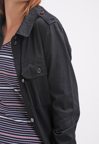 Veste en denim noir avec col boutonné, poches avant et épaulettes, portée par-dessus un haut rayé multicolore.