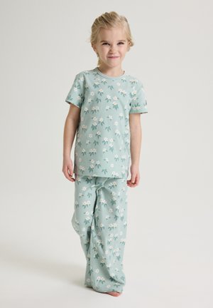 Jong kind in lichtgroene pyjama met bloemenpatroon, blootsvoets staand tegen een effen achtergrond.