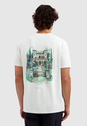 Man met krullend haar die een wit t-shirt draagt met een kleurrijke illustratie van een tuin en landhuis en de tekst "Droom groot, groter" op de achterkant.