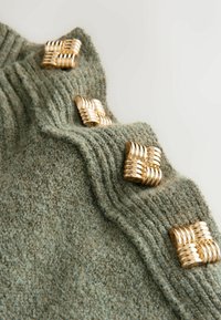 Grüner strukturierter Pullover mit geripptem Muster und goldenen metallischen Akzenten, die an gewebte Quadrate auf der Schulter erinnern. Aus weichem Wollmischmaterial.