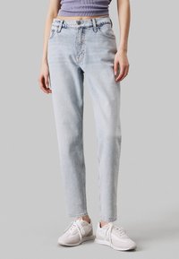 Jeans de denim azul claro con un diseño de pierna recta, cintura alta, un ligero desgaste y detalles cosidos, combinados con zapatillas blancas.