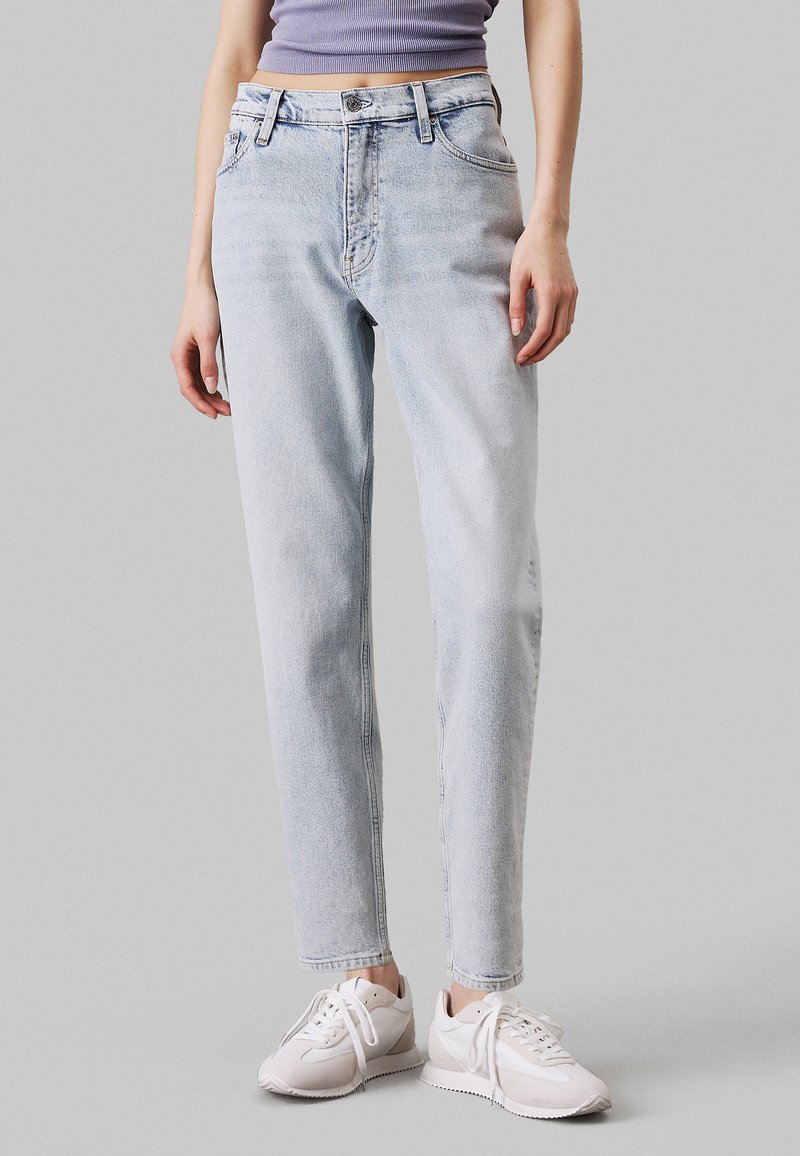 Jeans de denim azul claro con un diseño de pierna recta, cintura alta, un ligero desgaste y detalles cosidos, combinados con zapatillas blancas.