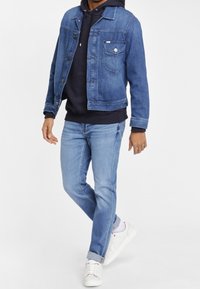 Giacca di jeans blu sopra un hoodie nero, abbinata a jeans blu chiaro e sneaker bianche. Presenta una tasca sul petto e dettagli con bottoni.