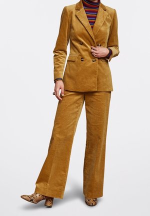 Persoon draagt een mosterdgele corduroy double-breasted blazer en bijpassende broek met enkellaarsjes met slangenprint en een gestreepte coltrui.