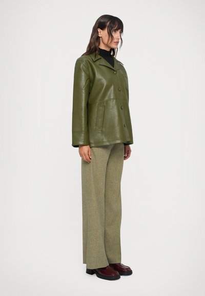 Giacca in ecopelle verde oliva con colletto a punta e bottoni frontali, abbinata a pantaloni larghi a fantasia verde chiaro e scarpe borgogna.