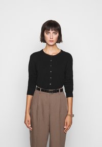 Cardigan nero a maglia con scollatura rotonda e chiusura con bottoni, abbinato a pantaloni beige a vita alta e una cintura nera. Accento di bracciale dorato.