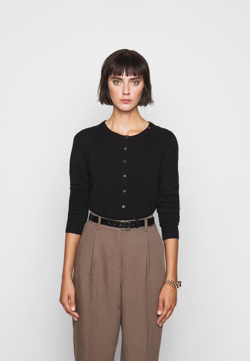 Cardigan nero a maglia con scollatura rotonda e chiusura con bottoni, abbinato a pantaloni beige a vita alta e una cintura nera. Accento di bracciale dorato.