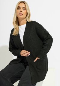 Svart, stickad cardigan med öppen framsida, långa ärmar, vriden textur och sidfickor, i kombination med en vit T-shirt och mörka byxor.