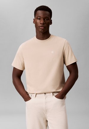 Jonge man met een donkere huid die een beige T-shirt met korte mouwen en bijpassende lichtbeige broek draagt, handen in de zakken, neutrale grijze achtergrond.