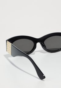 Black sunglasses with dark lenses, featuring a rounded frame and textured beige accents on the temples. Smooth plastic material, modern design.