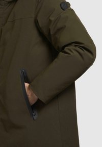 Green insulated jacket featuring a zippered pocket and a textured surface. A round black logo patch is visible on the shoulder.