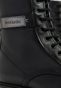 Zwarte leren enkelboot met een gladde textuur, ronde neus, metalen oogjes en een grijze merklabel aan de zijkant.