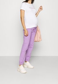 Tenue de maternité comprenant un t-shirt blanc ajusté avec un texte coloré, associé à un jogging lavande ample et des baskets blanches.