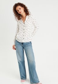 Chemise blanche à boutons avec un motif de cœurs noirs, manches longues et ourlet arrondi, associée à un jean bleu clair à jambes larges et des baskets roses.