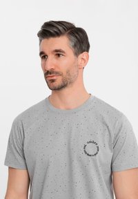 Camiseta de algodón gris con un logo negro redondo en el lado izquierdo del pecho, con motas negras dispersas por toda la tela. Cuello redondo, mangas cortas.
