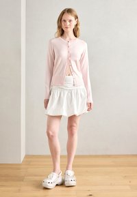 Cardigan rose en maille avec encolure ronde et fermeture à boutons, associé à une jupe plissée blanche. Sabots blancs ornés de fleurs.