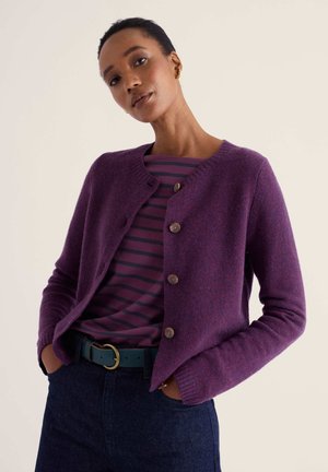 Femme portant un cardigan violet boutonné sur une chemise rayée violette et noire, avec un jean bleu foncé et une ceinture bleu sarcelle avec une boucle dorée.