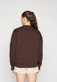 Sweatshirt marron en tissu doux, avec un col rond, des épaules tombantes et des poignets côtelés. Porté avec un short de couleur claire.