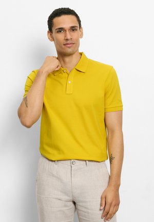 Mann trägt ein gelbes Poloshirt und hellbeige Hose, richtet mit der linken Hand den Hemdkragen, steht vor einem weißen Hintergrund.