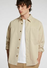 Camicia beige a maniche lunghe con chiusura a bottoni, taschino sul petto e texture di tessuto morbido, indossata sopra una maglietta bianca.