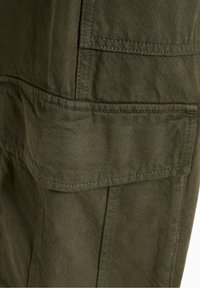 Gros plan sur une poche cargo en tissu vert olive avec rabat et coutures visibles sur un pantalon ou une veste.