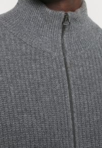 Grå stickad tröja med hög krage och dragkedja fram. Har en ribbad textur och en slät hårdvaruaccent nära dragkedjan.