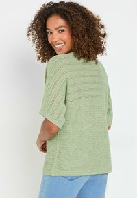 Maglione a maglia verde chiaro con una vestibilità ampia, dotato di maniche corte e un design a righe testurizzato. Indossato con jeans di denim blu.