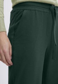 Dark green, elastic-waist pants with a smooth texture, featuring side pockets and a tie closure. Visible stitching detail at the waist.
