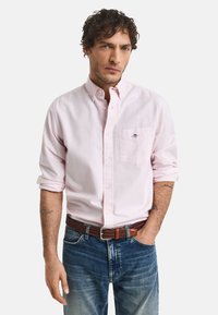 Ljusrosa skjorta med knappar, bröstficka, upprullade ärmar och en subtil logotyp, kombinerad med blå denimjeans och ett brunt flätat bälte.