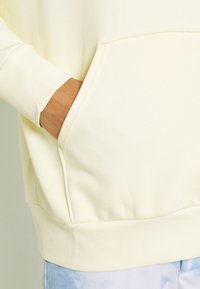 Felpa gialla chiara con polsini a costine e tasca frontale, caratterizzata da una consistenza morbida e una vestibilità leggermente oversize.