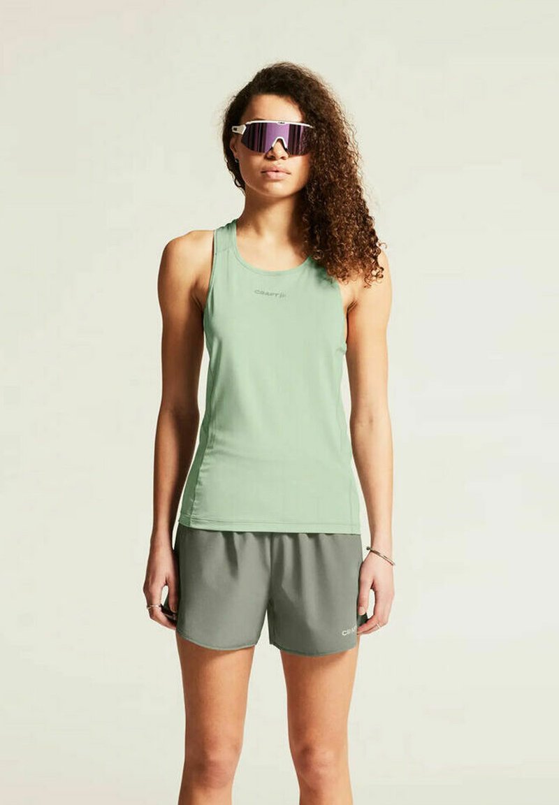Jonge vrouw met krullend haar, draag een reflecterende sportzonnebril, een mouwloze groene top en grijze sportshorts, staand tegen een effen achtergrond.