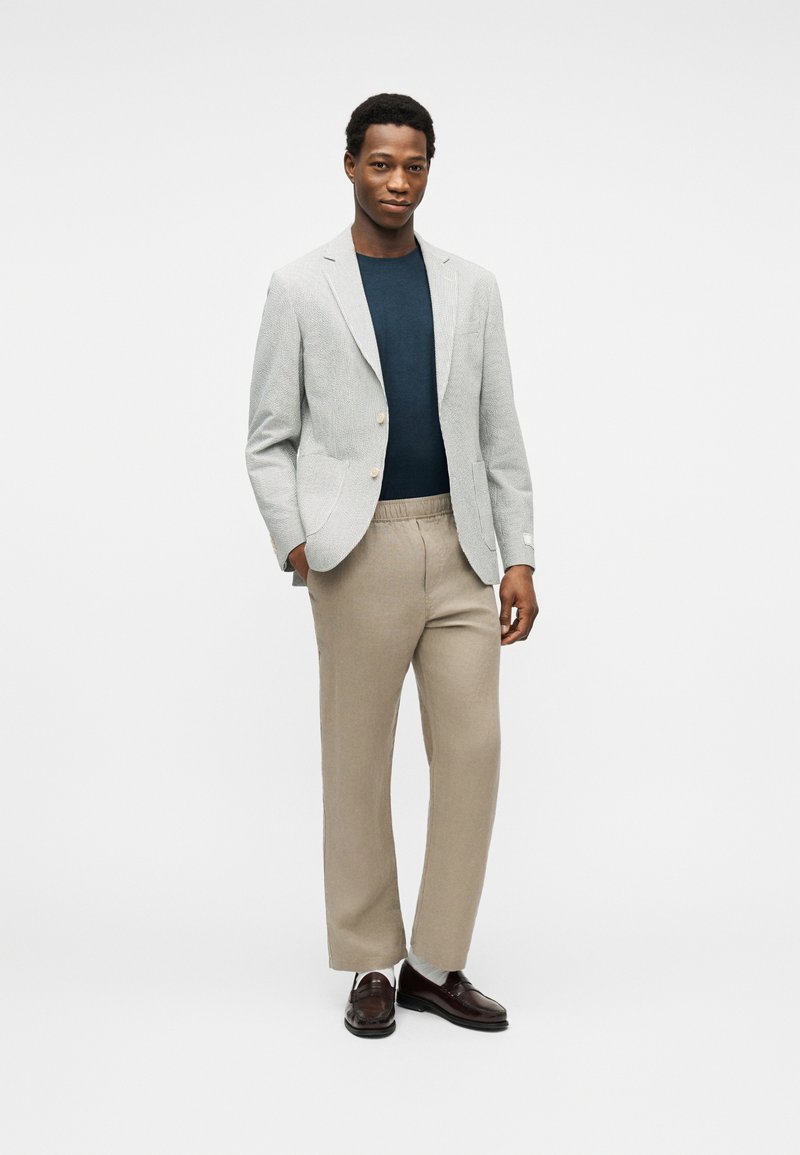 Uomo con blazer grigio chiaro, camicia blu scuro, pantaloni beige e mocassini marrone scuro, in piedi davanti a uno sfondo bianco.