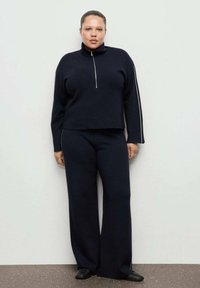 Temno modra pletena usklajena obleka z zadrgo na turtleneck in širokimi hlačami, s kontrastnimi belimi bočnimi črtami in mehko teksturo.