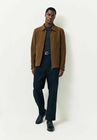 Mann in braunem Reißverschlussjacke, dunkelgrauem Pullover, schwarzem Gürtel, schwarzen Hosen und schwarzen Schuhen, der vor einem schlichten weißen Hintergrund steht.