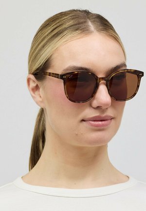 Vrouw met blond haar naar achteren gebonden, draagt een schildpadzonnebril en een wit shirt met ronde hals, kijkt iets naar rechts.