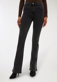 Kobieta ubrana w ciemnoszare jeansy typu bootcut z wysokim stanem oraz czarne szpilki, stoi z rękami swobodnie opuszczonymi przy bokach.