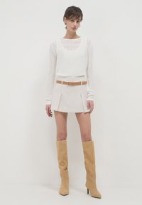 Weißes, strukturiertes Langarmoberteil, hellbeige, plissierter Minirock mit braunem Gürtel und knielangen, beigen Wildlederstiefeln. Minimalistisches Design, neutrale Farben.