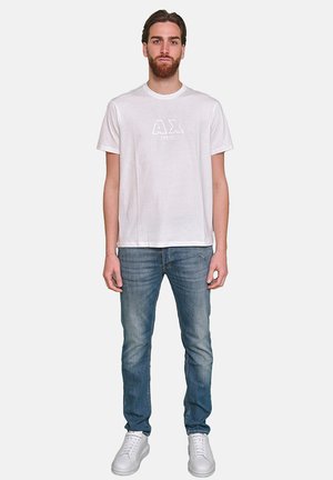 Maglietta bianca in cotone con collo rotondo e maniche corte, caratterizzata da un grande logo "AX" al centro. Indossata con jeans di denim blu e sneakers bianche.