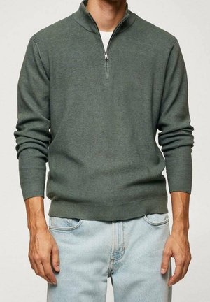 Dunkelgrüner, strukturierter Reißverschluss-Pullover mit Stehkragen und langen Ärmeln. Über einem weißen Shirt getragen, kombiniert mit hellblauen Jeans.