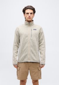 Νέος άνδρας που φοράει μπεζ φλις μπουφάν με φερμουάρ και ταμπά cargo σορτς, στέκεται μπροστά σε απλό λευκό φόντο.